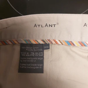 Beige byxor från Atlant - Säljer ett par stilrena beige byxor från Atlant. De är tillverkade i 100% bomull vilket gör dem både bekväma och hållbara. Byxorna har en klassisk rak passform och är perfekta för både vardag och mer formella tillfällen. De har en enkel design med knappar baktill och en färgglad randig detalj på insidan av midjan.