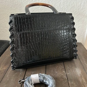 Svart handväska med krokodilmönster - Säljer en svart handväska med krokodilmönster. Den har snygga detaljer med snörning på sidorna och ett robust handtag i trä! Väskan är i nyskick och kommer med en avtagbar axelrem. Fri frakt