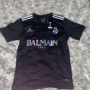 Svart fotbollströja från Adidas - Säljer en snygg svart fotbollströja från Adidas med coola detaljer. Tröjan har korta ärmar och ett holografiskt Balmain Paris-tryck på framsidan. Den har också Real Madrid-loggan och Adidas-loggan i färg. Perfekt för fotbollsfans som vill ha något unikt!