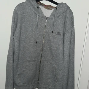 Grå hoodie med dragkedja - Säljer en snygg grå hoodie med dragkedja och fickor fram. Den har en cool rutigt mönstrad insida av huvan som ger en extra touch. Perfekt för höst och vinter, och den är superbekväm att ha på sig. Passar både till vardags och chillkvällar.