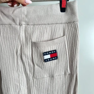 Beige ribbade byxor från Tommy Jeans - Säljer ett par snygga beige ribbade byxor från Tommy Jeans. De är högmidjade och har en skön, loose passform. Perfekta för en avslappnad stil men ändå trendiga. Byxorna är i nyskick och har en liten ficka med Tommy Jeans-loggan på framsidan. Superbekväma och lätta att matcha med olika toppar! köpt för 999kr