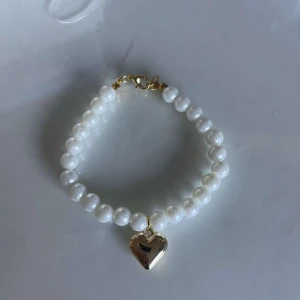 Pärlarmband med hjärthänge - Säljer ett superfint armband med vita pärlor och ett gulligt guldfärgat hjärthänge. Perfekt för att ge en romantisk touch till din outfit. Armbandet har en enkel och elegant design med ett praktiskt lås.