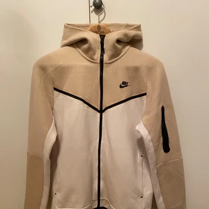 Nike Tech Fleece, beige och vit (M) - Säljer en snygg och sportig jacka från Nike i beige och vitt. Jackan har en dragkedja framtill och en huva för extra skydd. Perfekt för vår och höst!