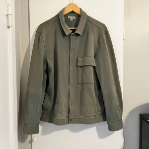 Grön overshirt från COS - Säljer en stilren grön overshirt från COS. Den har en klassisk krage och knappar framtill. Perfekt för lager-på-lager under hösten. Den har en praktisk bröstficka och är i ett mjukt material som känns bekvämt att bära.