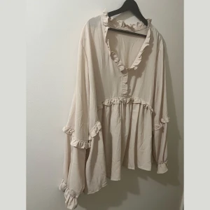 Beige blus med volangdetaljer - Säljer en superfin beige blus L-XL med volangdetaljer. Den har långa ärmar och en lös passform