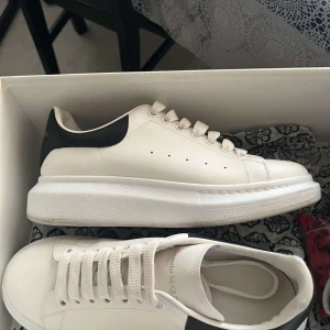 Vita sneakers från Alexander McQueen - Säljer ett par stilrena vita sneakers från Alexander McQueen. Skorna har en snygg svart detalj på hälen med märkesloggan i guld. De är i nyskick och kommer i originalförpackning. Perfekta för både vardag och fest!