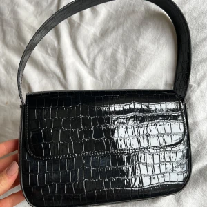 Svart axelväska i krokodilmönster - Snygg svart axelväska med krokodilmönster. Väskan har en stilren design med en enkel axelrem och stängs med en tryckknapp. Perfekt för både vardag och fest!