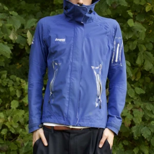 Blå vindjacka från Bergans - Vi Säljer en blå vindjacka från Bergans i bra skick. Jackan har en hög krage och flera praktiska dragkedjor på framsidan och ärmarna. Perfekt för höst- och vårväder. Den är långärmad och har en justerbar huva för extra skydd mot vinden.