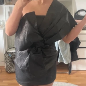 Svart glittrig klänning - Säljer en superfin svart glittrig klänning med en unik design. Den är axelbandslös och har en stor rosett framtill som ger en extra wow-effekt. Perfekt för festliga tillfällen! Klänningen är kort och har en dragkedja bak för enkel påklädning. Passar perfekt för en utekväll eller nyårsfest! ✨