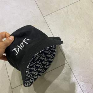 Säljer en snygg bucket hat från Dior. Den är svart med vit broderad logga och har ett coolt mönster på insidan. Perfekt för att ge din outfit en lyxig touch. Passar både till vardags och fest!