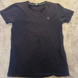 Svart t-shirt från GANT - Säljer en stilren svart t-shirt från GANT i storlek 146/152 cm, perfekt för åldrarna 11-12 år. T-shirten är i bomull och har en liten logga på bröstet. Den är kortärmad och passar bra till både vardag och fest. Skön och enkel att matcha med andra plagg!