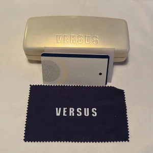 Versus Versace glasögonfodral - Snyggt glasögonfodral från Versus Versace i en stilren beige färg med en lyxig orange insida. Perfekt för att skydda dina glasögon med stil. Kommer med en rengöringsduk och autenticitetskort. Perfekt för dig som älskar mode och vill ha det lilla extra.