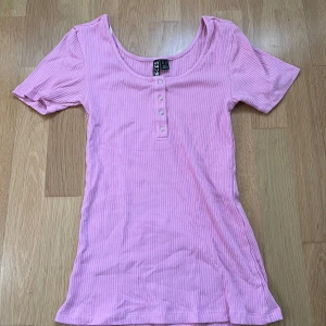 Rosa ribbad topp från Pieces - Säljer en superfin rosa ribbad topp från Pieces. Den har korta ärmar och en rund halsringning med knappar framtill. Använd ca 2 gånger. Står storlek M men den passar som XS