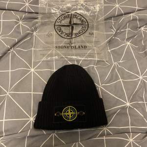 Säljer en snygg svart stickad mössa från Stone Island. Den är ribbad och har en cool logga framtill. Perfekt för att hålla sig varm och stilren under kalla dagar. Kommer med originalförpackning.