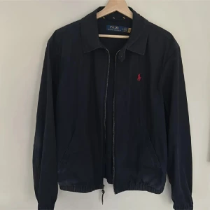 Marinblå jacka från Polo Ralph Lauren - Säljer en stilren marinblå jacka från Polo Ralph Lauren med dragkedja och klassisk krage. Jackan har en röd broderad logotyp på bröstet och elastisk nederkant för en bekväm passform. Nypris: 1999kr 