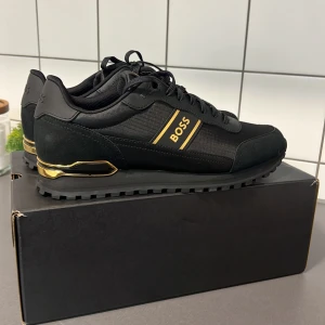 Svarta sneakers från Boss - Snygga svarta sneakers från Boss med gulddetaljer och logotyp på sidan. Skorna har snörning och en robust sula för extra grepp. Perfekta för en stilren look. Det som tillkommer är sko boxen till skorna. Ny pris 2400 Pris kan diskuteras vid en snabb affär