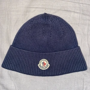Mörkblå stickad mössa från Moncler - Snygg mörkblå stickad mössa från Moncler med uppvikt kant och broderad logotyp framtill. Perfekt för kyliga dagar och en stilren look.