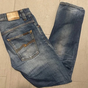 Nudie jeans - Säljer ett par snygga blå jeans från Nudie med klassisk femficksdesign och orange sömmar. Jeansen har en straight passform och är tillverkade i slitstarkt denim. Perfekta för en avslappnad stil.