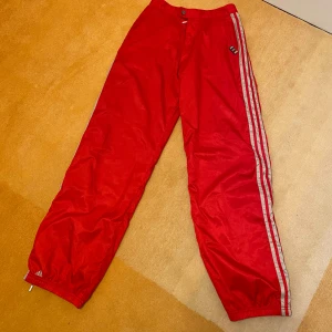 Röda träningsbyxor från Adidas - Retro röda träningsbyxor från Adidas med vita ränder längs sidorna. Byxorna har elastisk midja och dragkedja vid bensluten för extra komfort. Storlek XS eller 170. Mycket bra skick!