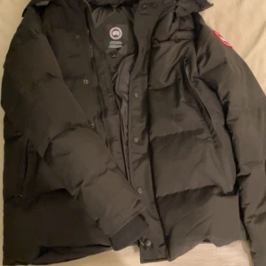 Svart dunjacka från Canada Goose - Säljer en svart dunjacka från Canada Goose med huva och knappar. Jackan har en klassisk design med varumärkets logga på ärmen. Perfekt för kalla vinterdagar.