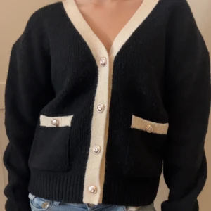 Svart kofta med vita detaljer - Säljer en snygg svart kofta med vita kanter och knappar. Koftan har två fickor fram och långa ärmar. Perfekt för en stilren look.