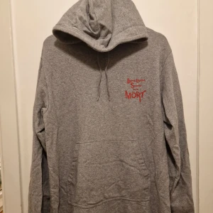 Grå hoodie från Cheap Monday - Säljer en grå hoodie från Cheap Monday med tryck i rött på bröstet. Tröjan har en klassisk känguruficka och justerbar huva med dragsko. Perfekt för en avslappnad stil. Garderobsrensning så kika gärna på övriga auktioner.