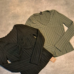 Ribbad tröja i svart och grön - Säljer två ribbade tröjor i svart och grön. Båda har en v-ringad design med långa ärmar och en snygg rynkdetalj framtill. Perfekta för en stilren look.