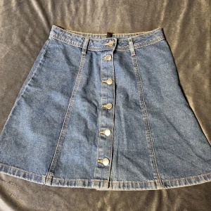 Jeanskjol från H&M Divided - Snygg jeanskjol från H&M Divided med knappar framtill. Kjolen har en klassisk A-linjeform och bälteshällor i midjan. Perfekt för en avslappnad stil.