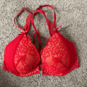 Röd spets-bh med rosett hunkemöller - Snygg röd bh i spets med dekorativ rosett och justerbara axelband. Den har en praktisk framknäppning. Den är oanvänd!