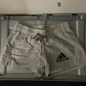 Grå shorts från Adidas - Säljer ett par grå shorts från Adidas i mjukt material. De har en elastisk midja med snörning och Adidas-loggan på ena benet. Perfekta för en avslappnad stil.