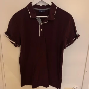 Vinröd pikétröja från Gant - Säljer en stilren mörkröd pikétröja från Gant med vita detaljer och knappar framtill. Tröjan har korta ärmar och en klassisk krage. Storlek L