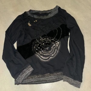 Svart långärmad tröja med unikt mönster - Säljer en svart långärmad tröja med ett unikt abstrakt mönster i silver och svart. Tröjan har dekorativa detaljer och ribbade kanter vid hals och ärmslut. Perfekt för dig som vill ha något annorlunda i garderoben!