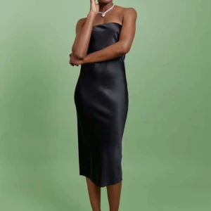 Svart axelbandslös klänning från Gina Tricot  - Elegant svart axelbandslös klänning i satin med en stilren design. Klänningen har en knytning baktill och en smickrande passform som passar perfekt för festliga tillfällen. Från Gina Tricot 