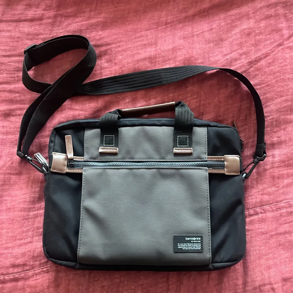 Snygg och praktisk datorväska från Samsonite i svart och grått. Väskan har en justerbar axelrem och flera fack med dragkedja för enkel organisering. Perfekt för att bära din laptop och andra viktiga tillbehör. Stilren design med detaljer i brunt läder. Man kan även sätta fast datorväskan på en resväska. Skriv vid frågor!. Laukut & Käsilaukut.