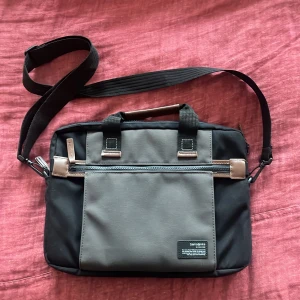 Svart och grå datorväska från Samsonite - Snygg och praktisk datorväska från Samsonite i svart och grått. Väskan har en justerbar axelrem och flera fack med dragkedja för enkel organisering. Perfekt för att bära din laptop och andra viktiga tillbehör. Stilren design med detaljer i brunt läder. Man kan även sätta fast datorväskan på en resväska. Skriv vid frågor!