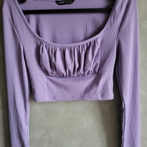 Lila ribbad topp från SHEIN - Snygg lila ribbad topp från SHEIN med långa ärmar och en croppad design. Toppen har en rundad halsringning och rynkade detaljer framtill som ger en fin touch. Perfekt för att styla med höga jeans eller kjol.