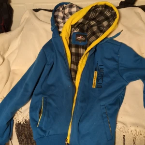 Blå zuoqi  jacka med dubbla huvor med gul dragkedja från Zuoqi - Säljer blå dubbel- jacka från Zuoqi med gul dragkedja och detaljer. Jackan har en fodrad insida med rutigt mönster och en huva. Perfekt för kyligare dagar. Aldrig använd prislappen kvar