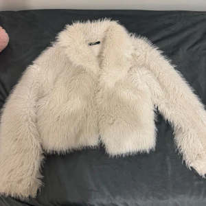 Vit pälsjacka från Gina Tricot - Säljer en fluffig och trendig vit pälsjacka från Gina Tricot. Jackan har en croppad design och långa ärmar, perfekt för att hålla sig varm och stilren under kyliga dagar. Ett måste för garderoben!