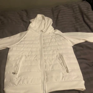 Vit cardigan  med dragkedja - Säljer en stilren vit cardigan med dragkedja och huva som aldrig är använd. Jackan har praktiska fickor med dragkedjor och är perfekt för kyligare dagar. Den quiltade designen ger en modern touch. Perfekt för dig som är 176 till 180 