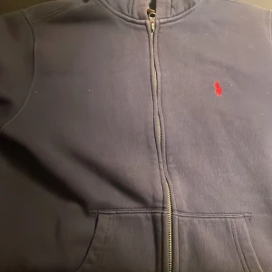 Mörkblå hoodie från Ralph Lauren - Säljer en snygg mörkblå hoodie från Ralph Lauren med dragkedja och röd broderad logga på bröstet. Tröjan har långa ärmar och en praktisk ficka framtill. Perfekt för en avslappnad stil. Pris skulle kunna diskuteras var inte rädd att fråga. 😄