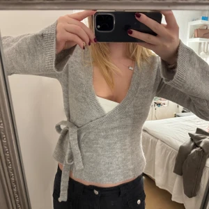 Grå omlottkofta med knytband - Säljer en stilren grå omlottkofta från ginatricot i storlek S. Koftan har långa ärmar och ett knytband i midjan för en justerbar passform. 