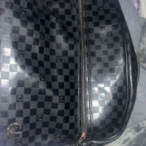 Svart rutig väska med dragkedja - Snygg svart väska med ett klassiskt rutigt mönster. Väskan har en praktisk dragkedja och är perfekt för att bära med sig det nödvändigaste. Den har en stilren design som passar till många tillfällen.