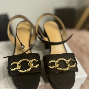 Svarta sandaler med gulddetaljer - Eleganta svarta sandaler med bred klack och guldfärgade kedjedetaljer framtill. Sandalerna har en justerbar rem med guldfärgat spänne runt ankeln. Perfekta för en stilfull look.