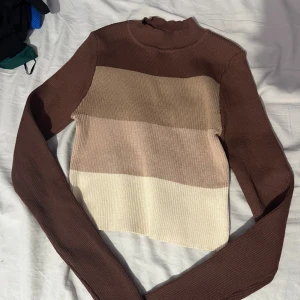 Randig stickad topp - Säljer en mysig stickad topp med breda ränder i brunt, beige och krämvitt. Den har långa ärmar och en hög hals.Passar utmärkt till både jeans och kjol.