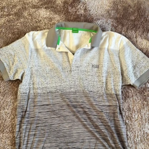 Grå och vit t-shirt från Boss - Snygg grå och vit t-shirt från Boss med korta ärmar och en klassisk krage. T-shirten har ett subtilt mönster och en diskret logga på bröstet. Perfekt för en stilren look.