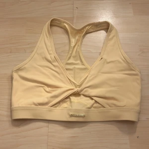 Gul sporttopp från Jessica Simpson - Snygg gul sporttopp från Jessica Simpson med knytdetalj framtill. Toppen har en brottarrygg och är tillverkad i ett mjukt och stretchigt material, perfekt för träning. Färgen tas inte riktigt upp i kameran, toppen är mer gul än vad det ser ut som på bild, lappen bortklippt storlek S/M