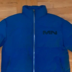 Blå pufferjacka från Mennace - Snygg blå pufferjacka från Mennace med dragkedja framtill. Jackan har en hög krage och ett stilrent tryck på bröstet och ryggen. Perfekt för kyliga dagar.