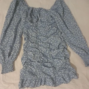 Blommig sommar klänning - Charmig blå klänning med små vita blommor och långa ärmar. Klänningen har en volangdetalj och en smickrande passform. Perfekt för en somrig look.