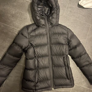 Svart dunjacka från Everest - Säljer en svart dunjacka från Everest med huva och dragkedja. Jackan är perfekt för kalla dagar och har en stilren design. Den är vadderad för extra värme och komfort.