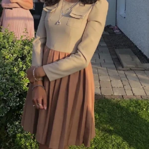 Vintage Beige klänning  - Elegant beige klänning från Louche med långa ärmar och dekorativa knappar på axlarna. Klänningen har en rund hals och två fickdetaljer på bröstet. Kjoldelen är plisserad och ger en fin volym.